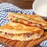 A ham and cheese quesadilla has been sliced and is ready to eat, served on a wooden board with a side of dip!