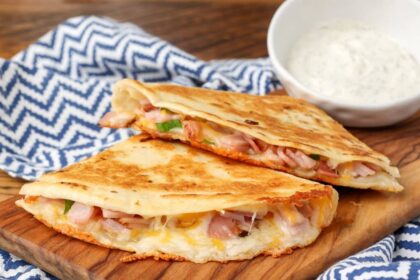 A ham and cheese quesadilla has been sliced and is ready to eat, served on a wooden board with a side of dip!